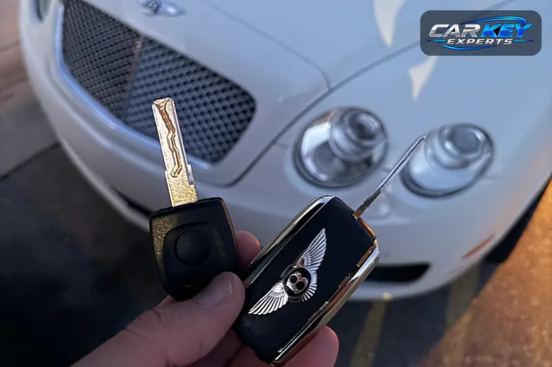 A new chip key and the original Bentley flip key held up in front of the white car in Nampa